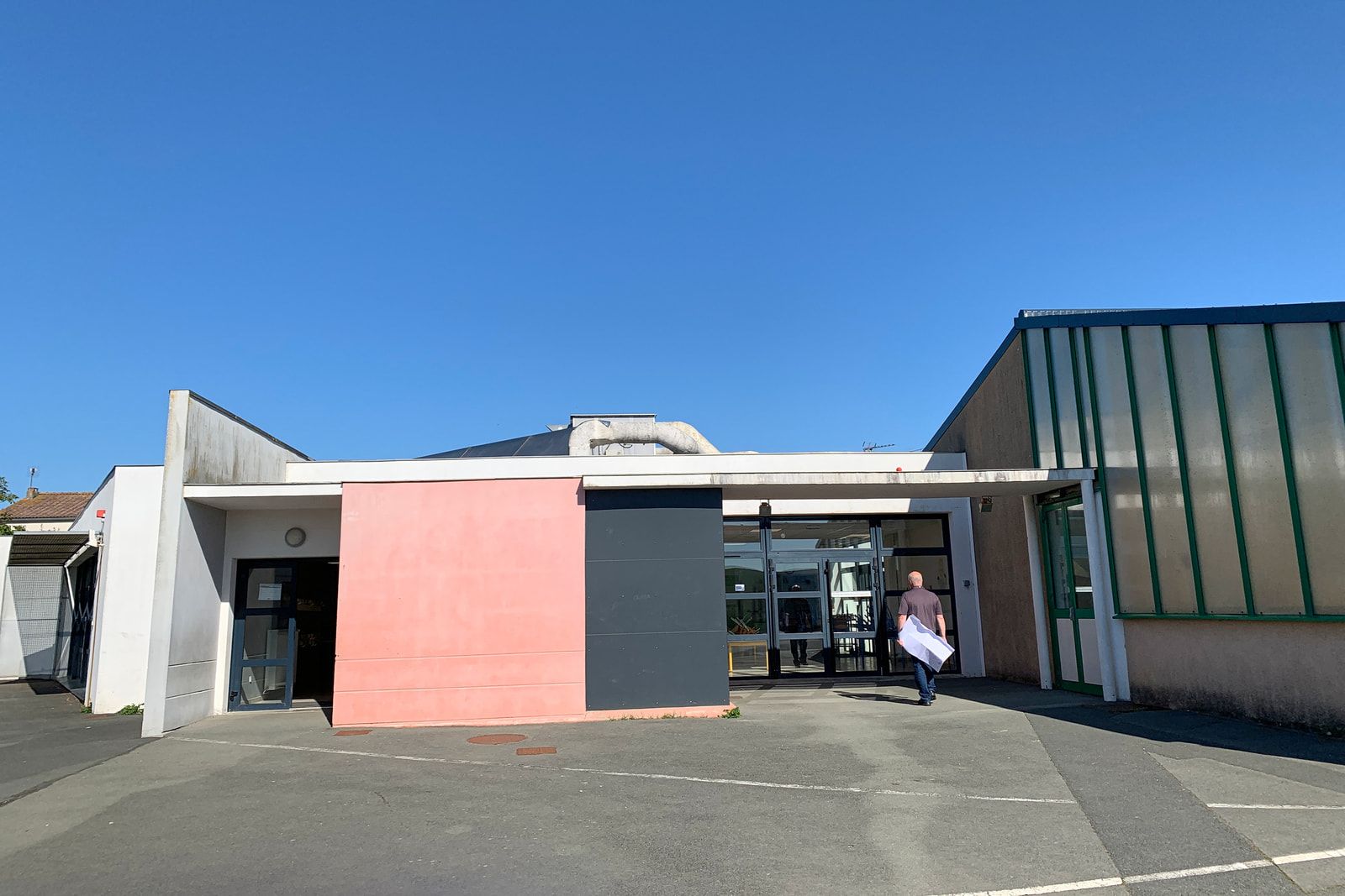 Entrée du restaurant scolaire de Villedoux sous auvent, entre un pan enduit rose et une façade à panneaux translucides.