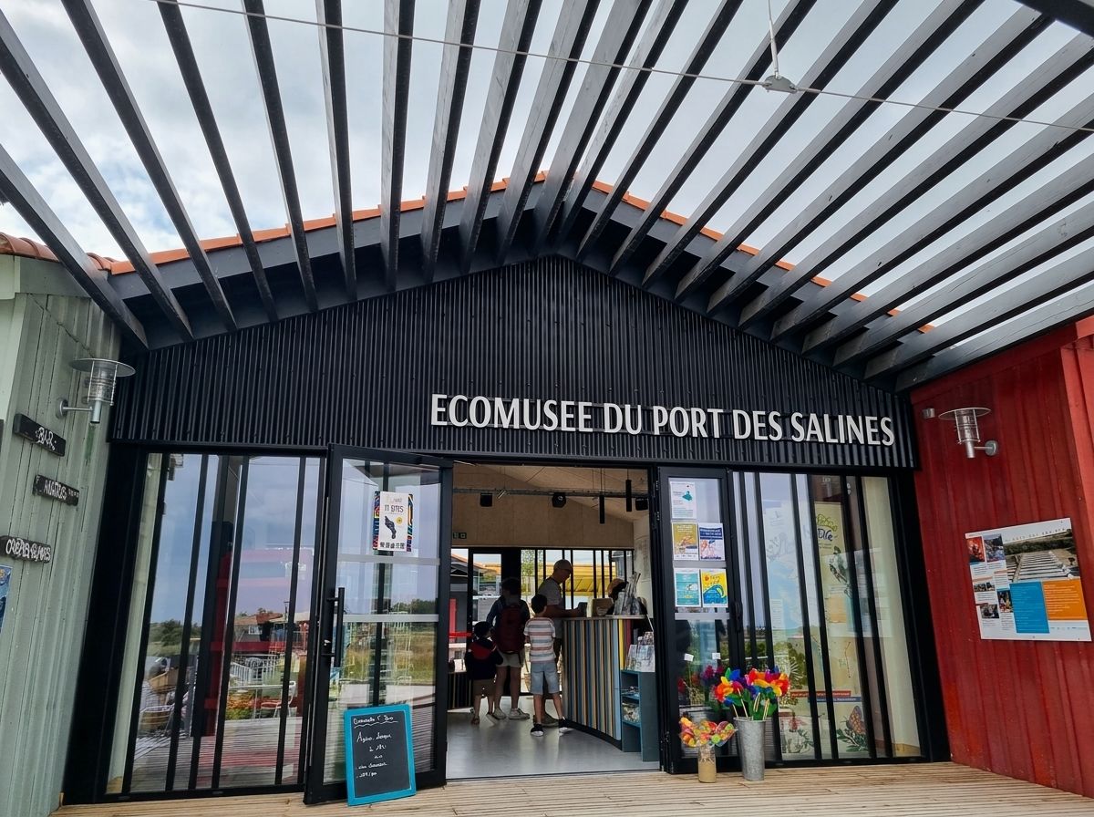 Entrée de l’Écomusée du Port des Salines sous un auvent à lames noires, entre deux cabanes à bardage coloré.